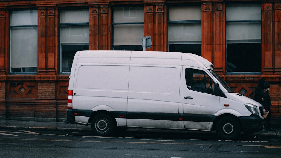 Two men engaged in a home relocation process outside a residential property, loading a large, rectangular cardboard box onto a hand truck. The men are dressed in casual clothing, with one wearing a ca