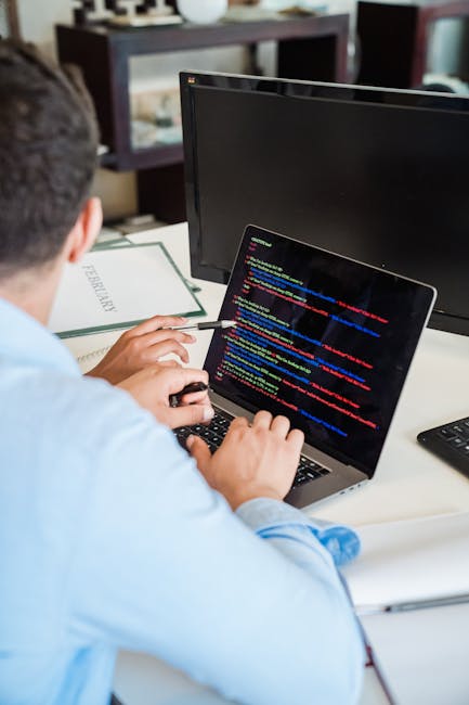 A person seated at a desk, working on a laptop displaying a screen with colorful lines of code, with a desktop monitor positioned behind the laptop and a black remote control on the right side of the 